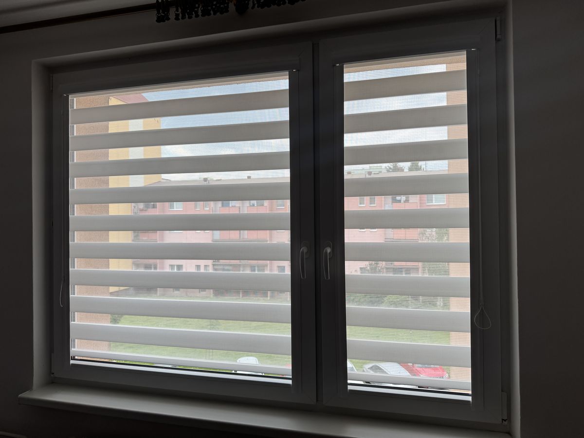 Rolety den a noc ve dvoukřídlém okně s tmavým rámem, výhled do ulice